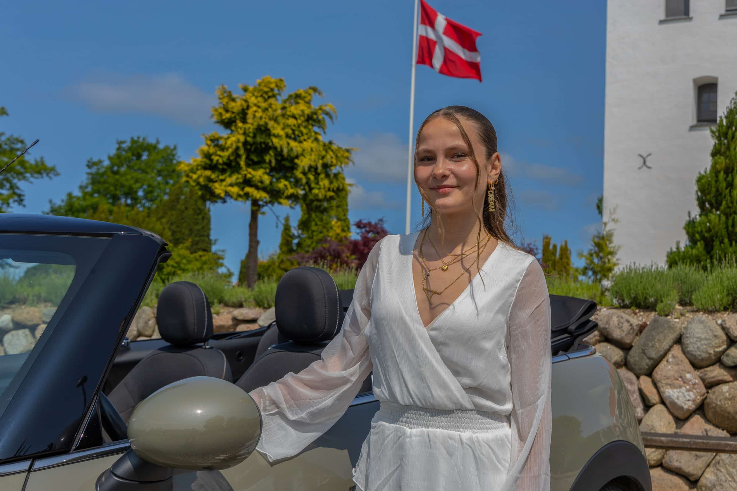 Kvindelig konfirmand står lænet op af sportsvogn med kirke og dannebrogsflag i baggrunden