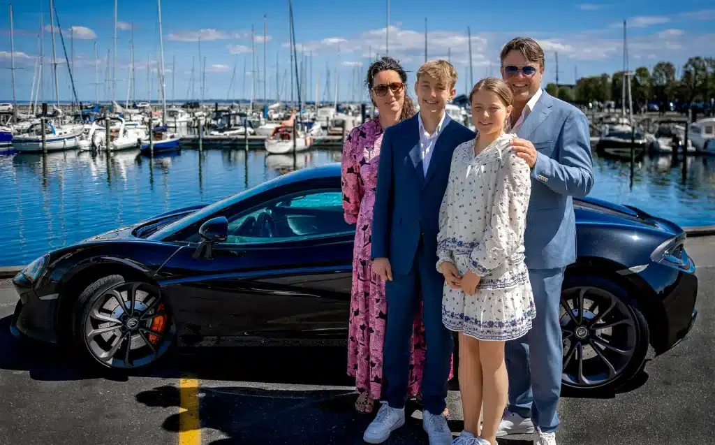 Konfirmations-fotograferne har her fotograferet en Konfirmand, der står sammen med sine forældre og søster med en sort sportsvogn bag sig og med havnen i baggrunden